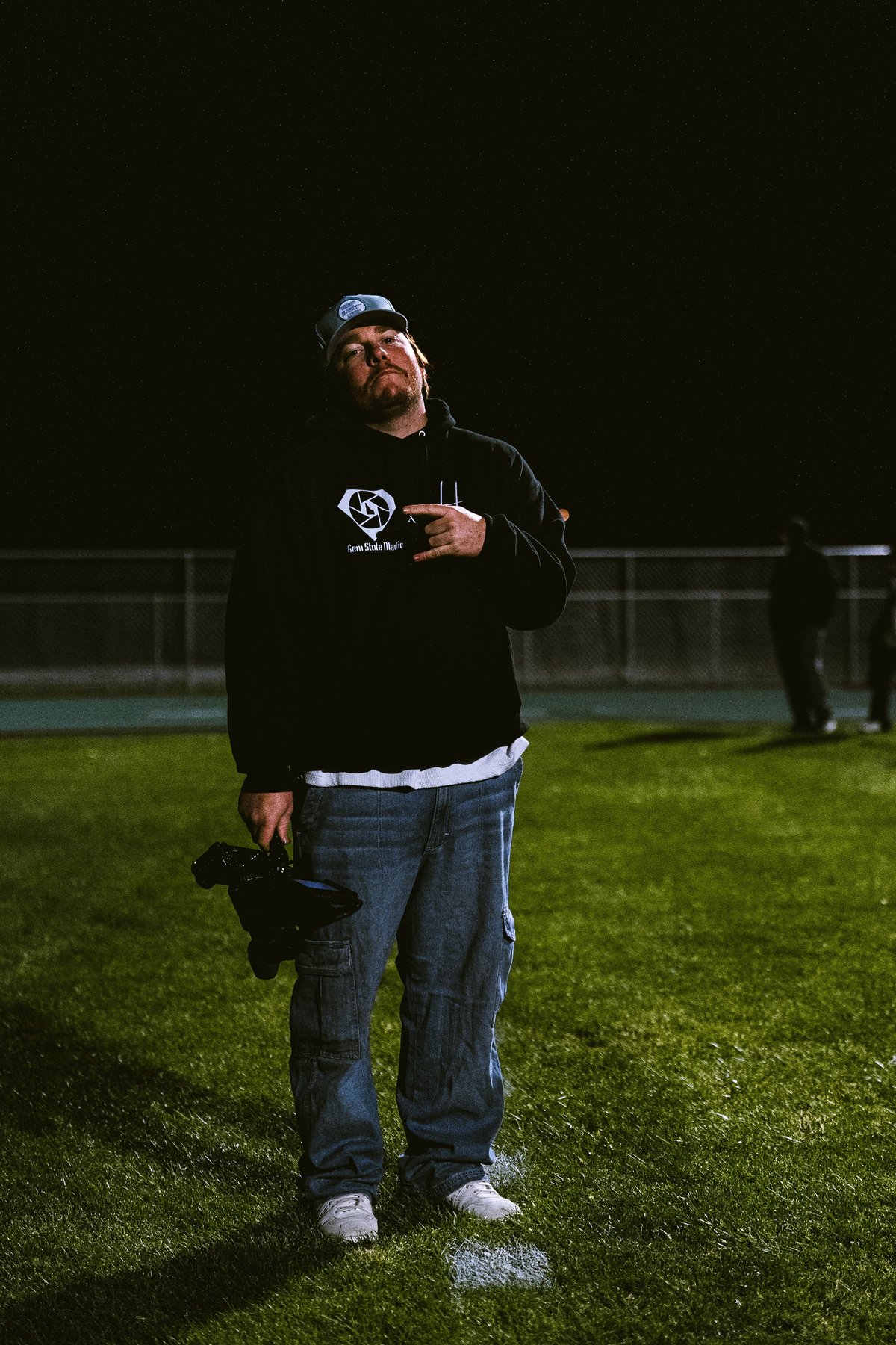 Man wearing black hoodie and blue jeans standing on a grass field at night holding a camera
